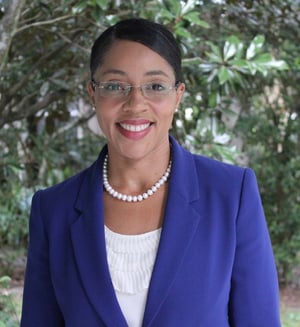 Headshot of smiling woman in front of green trees wearing a blue blazer, pearl accessories, and glasses.