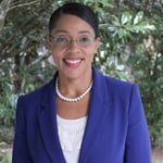 Headshot of smiling woman in front of green trees wearing a blue blazer, pearl accessories, and glasses.