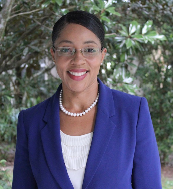 Headshot of smiling woman in front of green trees wearing a blue blazer, pearl accessories, and glasses.