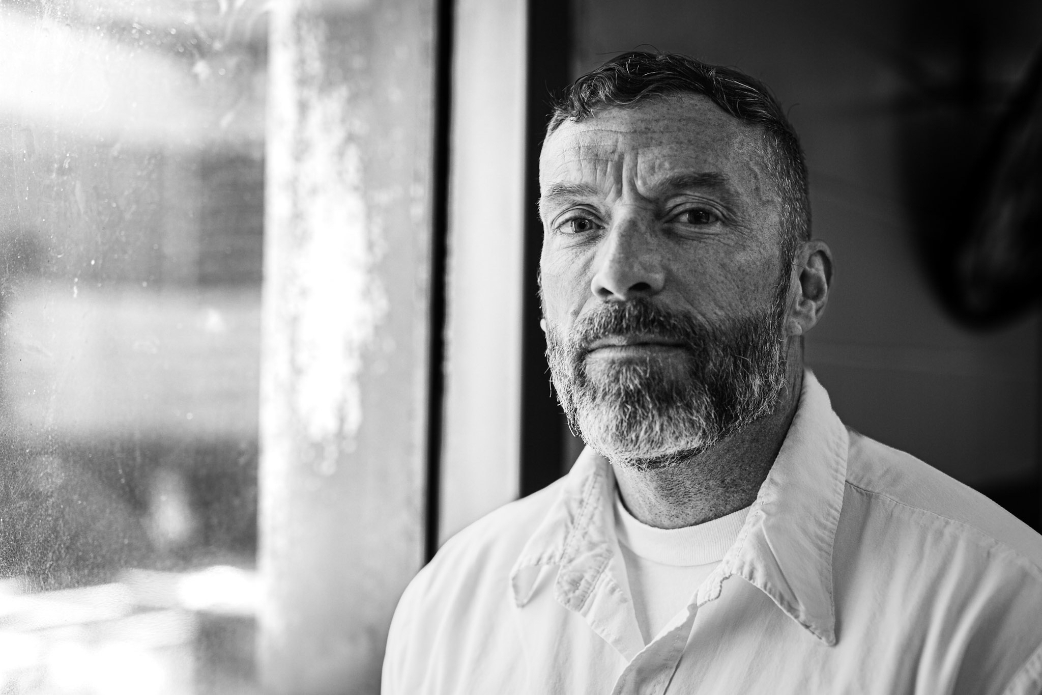 Middle-aged white man with short hair and graying beard next to window, in B&W