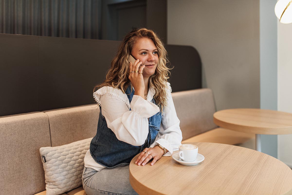 A woman with curly hair sitting on a grey sofa by a wooden table, talking on the phone.