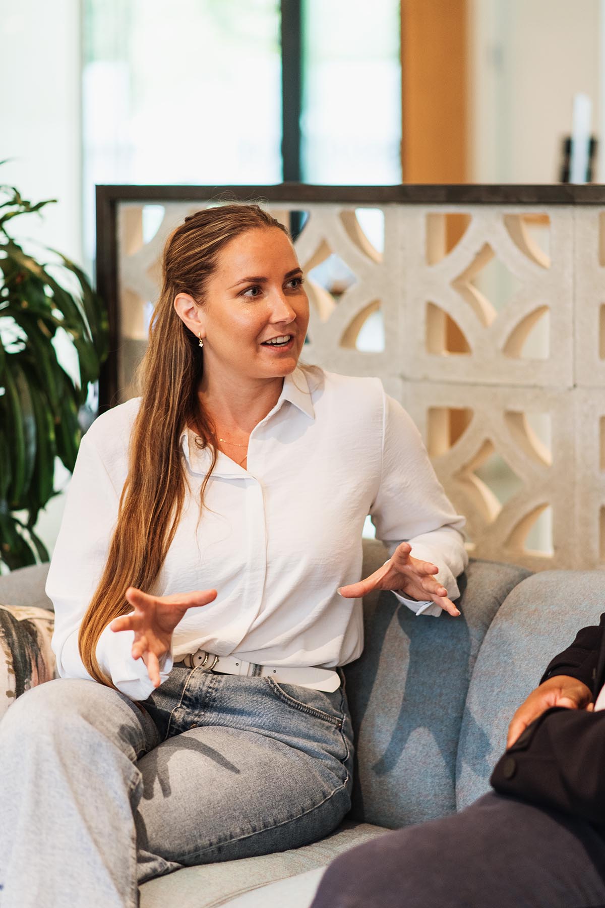 A woman in a white shirt talking to another person, only partially visible from the torso and leg, in a modern office environment.