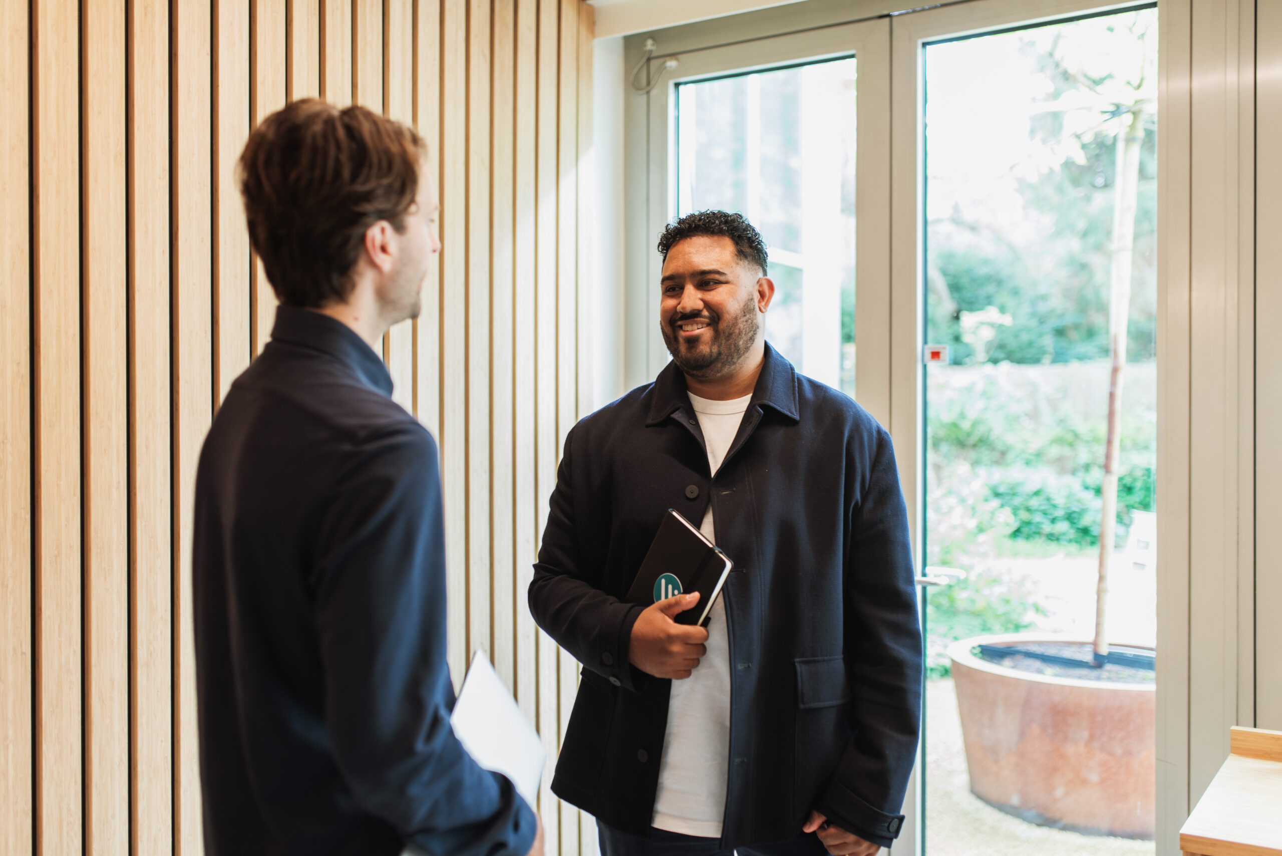 Two male colleagues stand in an office corridor discussing employment checks.
