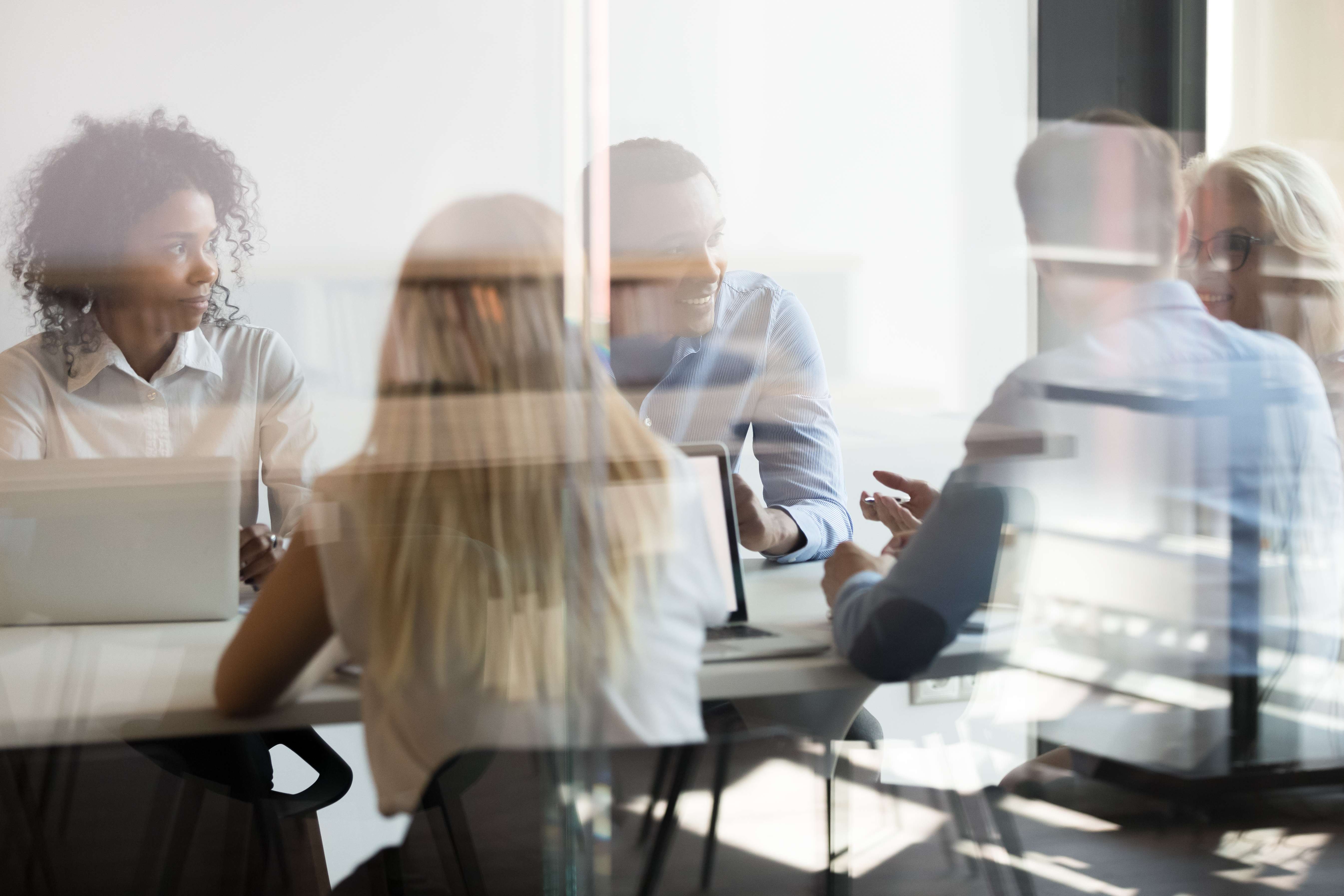 A group of people sitting around a table in an office environment, seen through a glass partition with subtle reflections on the glass, giving a sense of depth and modern workspace.