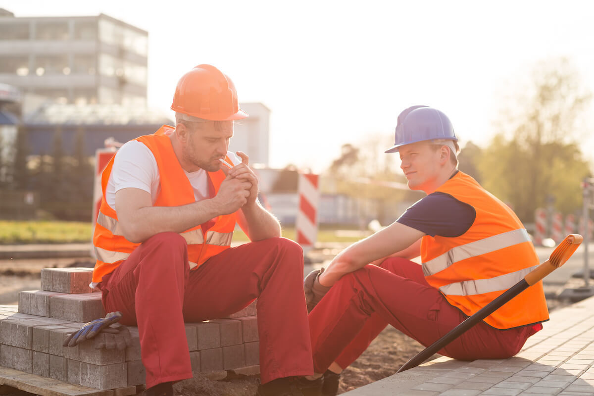 two men in hardhats and safety vests sit down for a cigarette