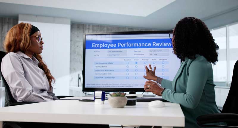 Employee Performance Review, an employee sits at a desk with their manager reviewing their performance on a computer screen.