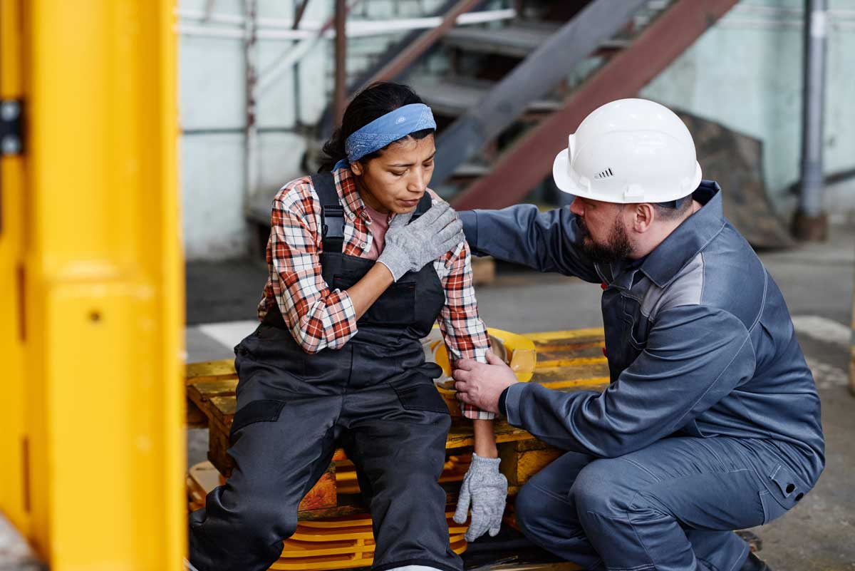 Foreman in workwear giving first aid to female worker with pain in her shoulder after accident at work