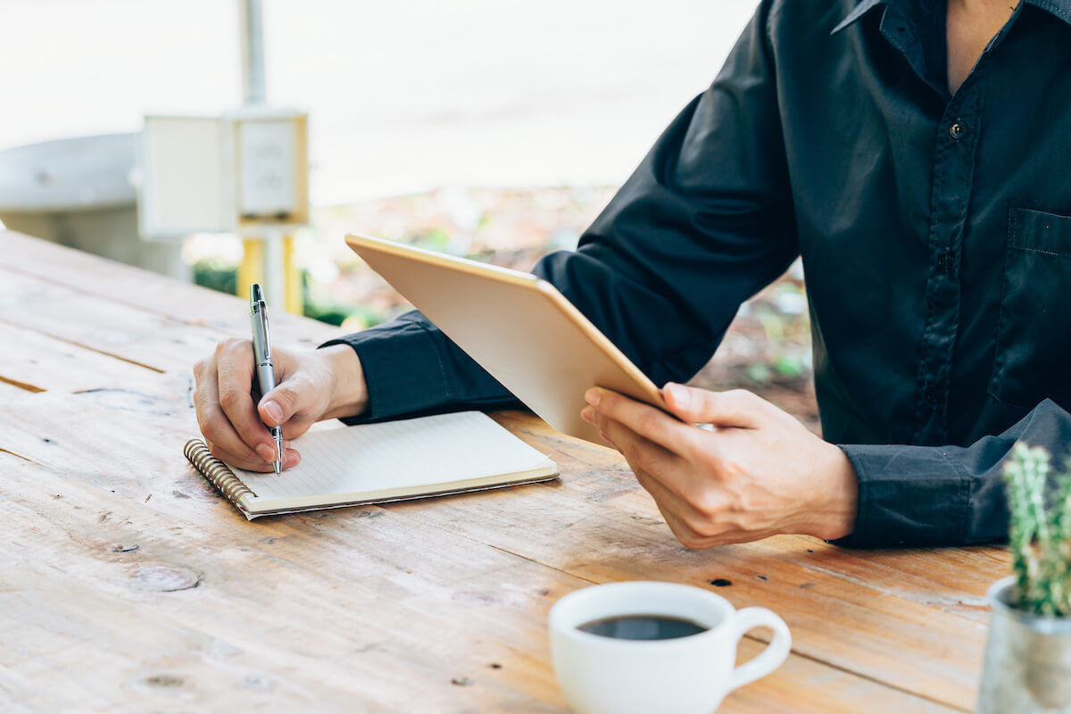 person taking notes on paper while holding tablet