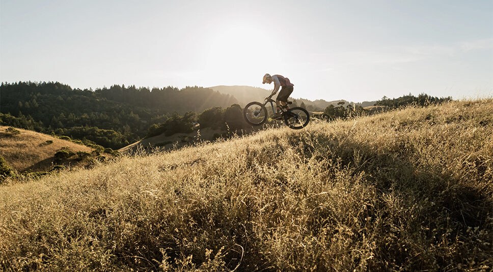 Person photographed in midair mountain biking down a hill with the sun low in the sky