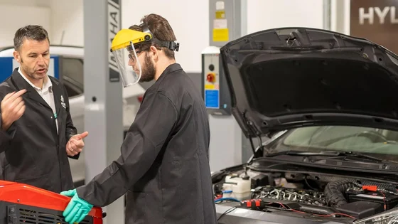 An assessor talking with an Hyundai apprentice with a Hyundai car in the background with its bonnet open inside a workshop.