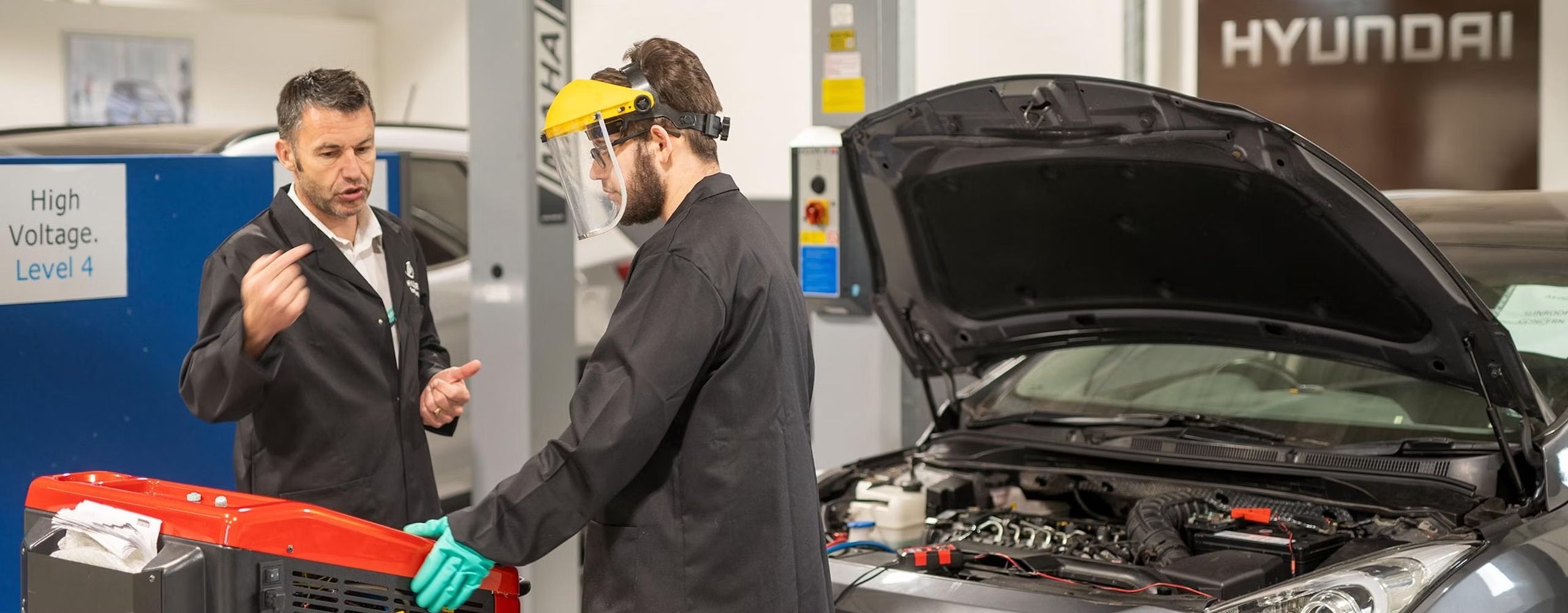 An assessor talking with an Hyundai apprentice with a Hyundai car in the background with its bonnet open inside a workshop.