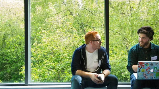 Two students sat talking to each other in front of big windows showing a woodland outside