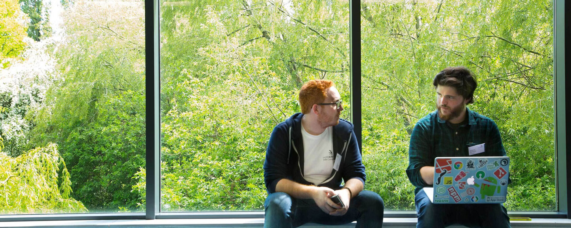 Two students sat talking to each other in front of big windows showing a woodland outside