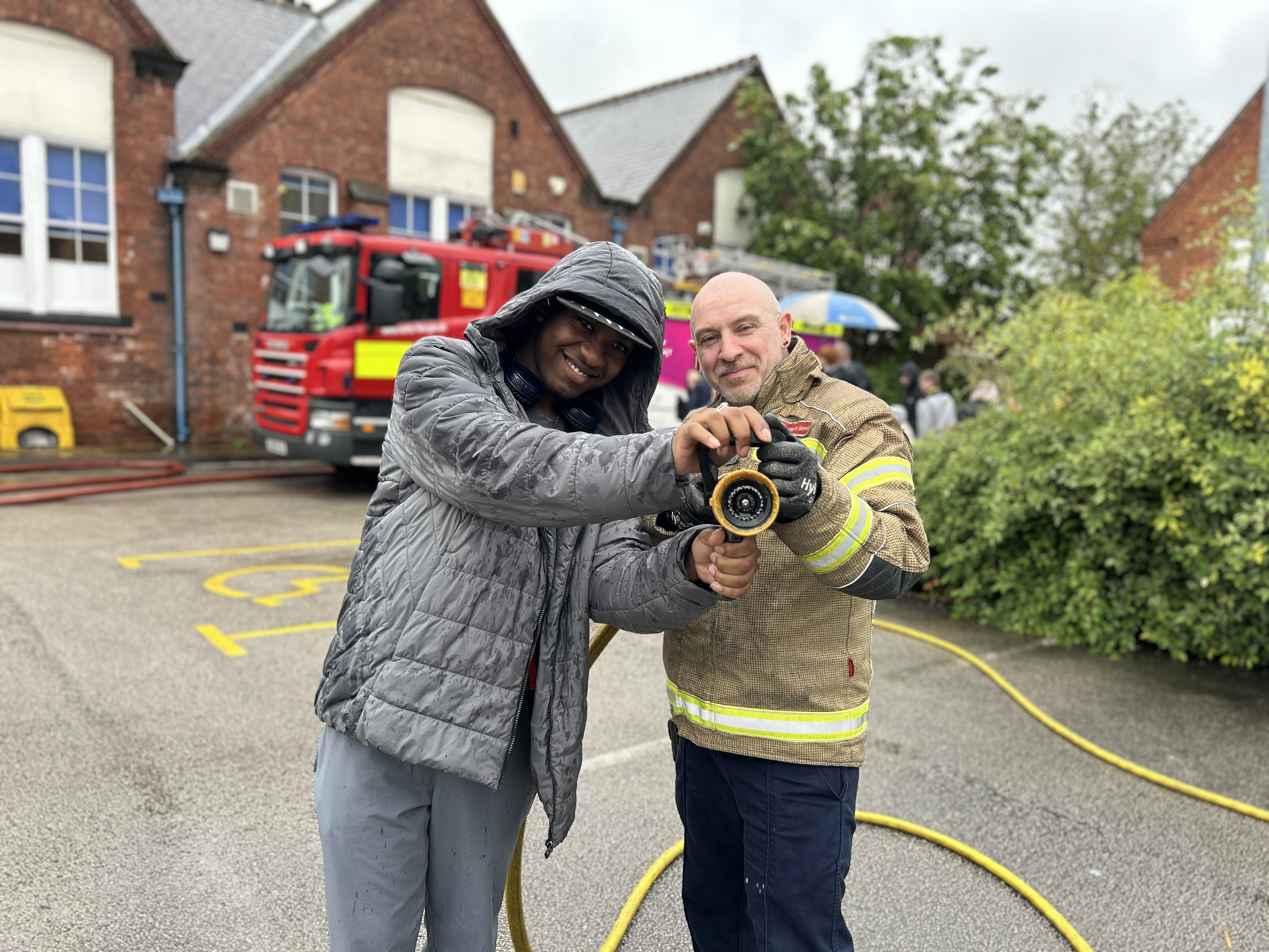 A student holding a hose with a firefighter standing in front of a fire engine.