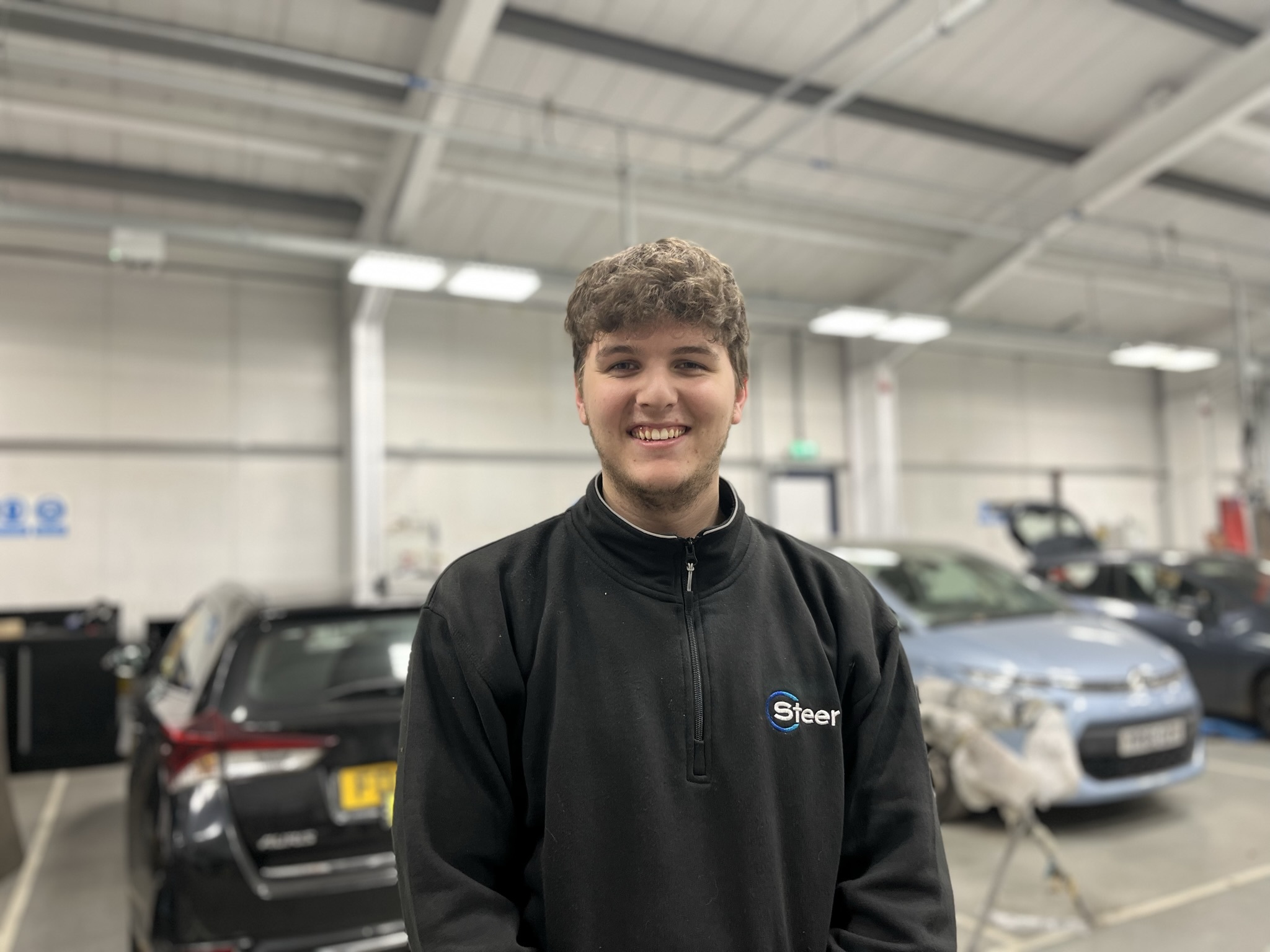 A student in an automotive workshop with cars behind him.