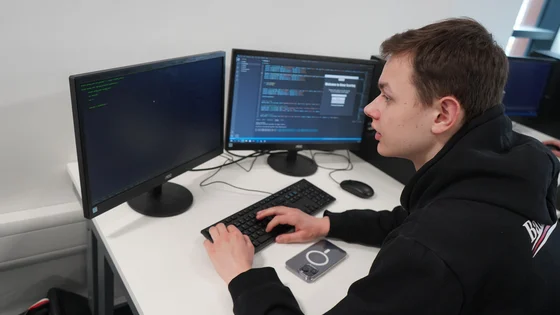 A student sitting at a desk with two screens showing C# code in Visual Studio.