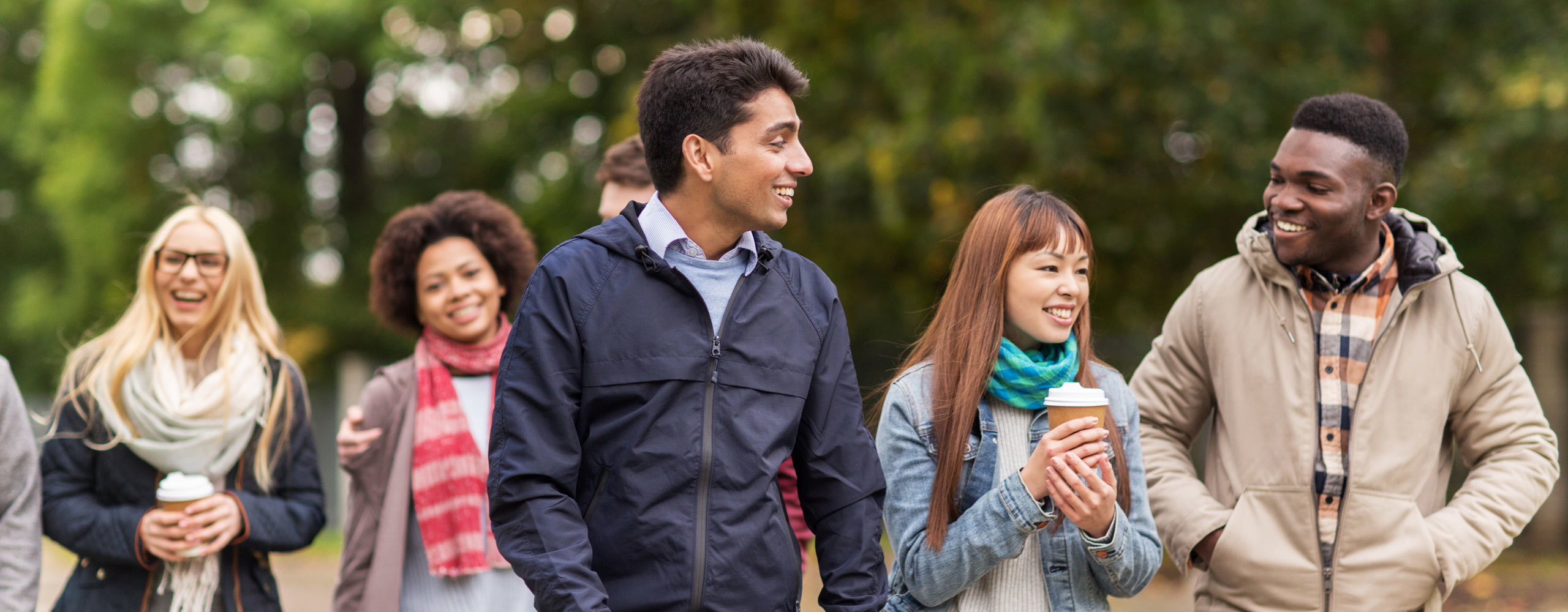 A group of young students walking in a park wearing scarves and carrying coffee.