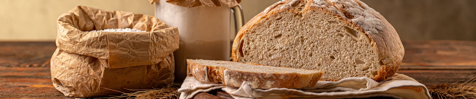 A hessian bag of flour, a jar with sourdough starter and a loaf of sourdough bread that has been cut.