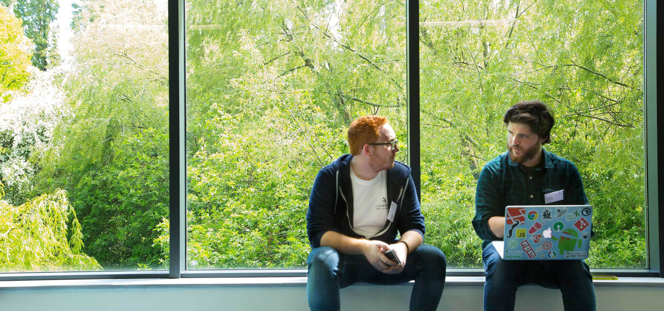 Two students sitting on a ledge, with a laptop with stickers