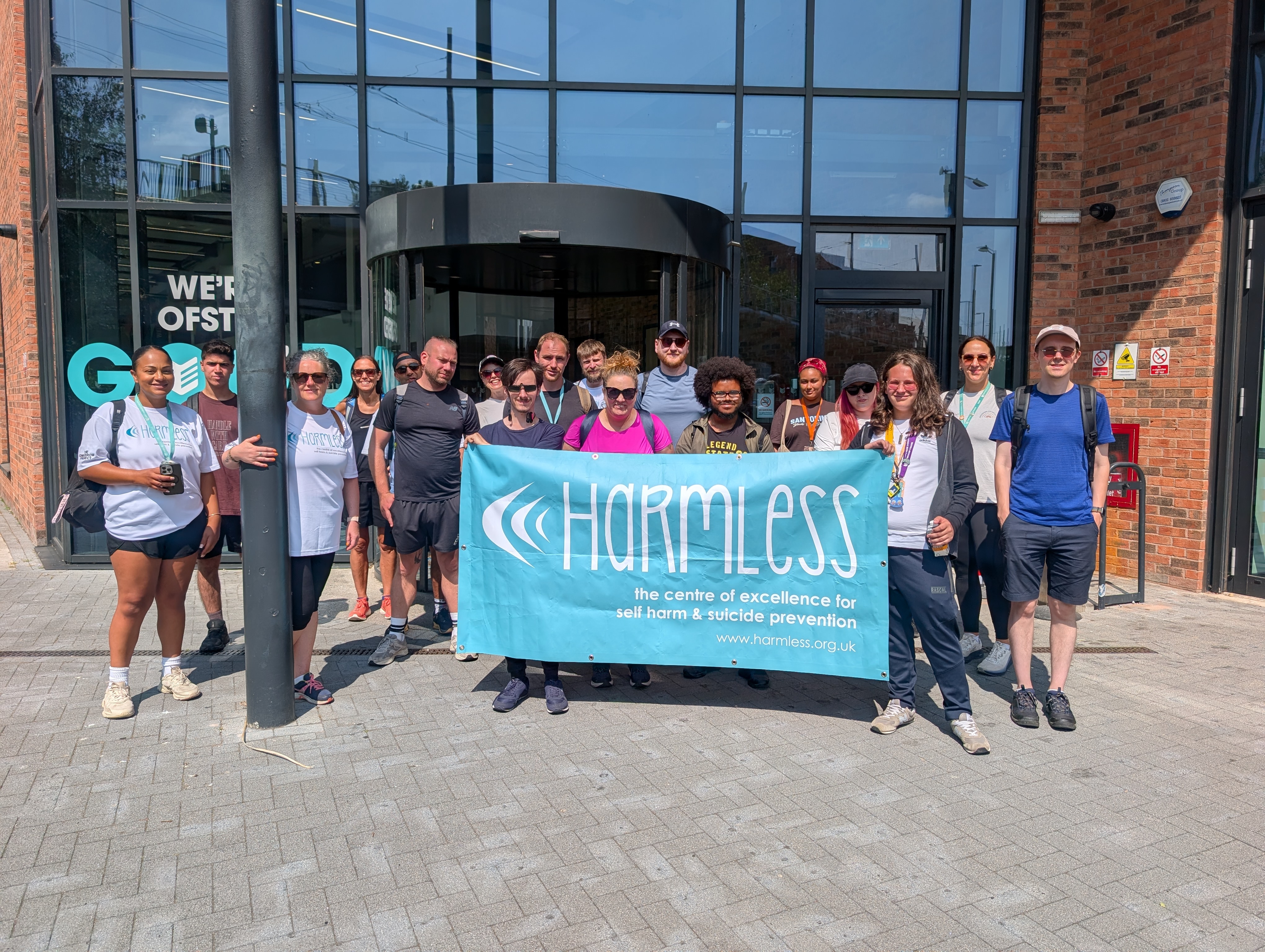 Staff and students outside City Hub holding a banner that has the charity Harmless text written on it