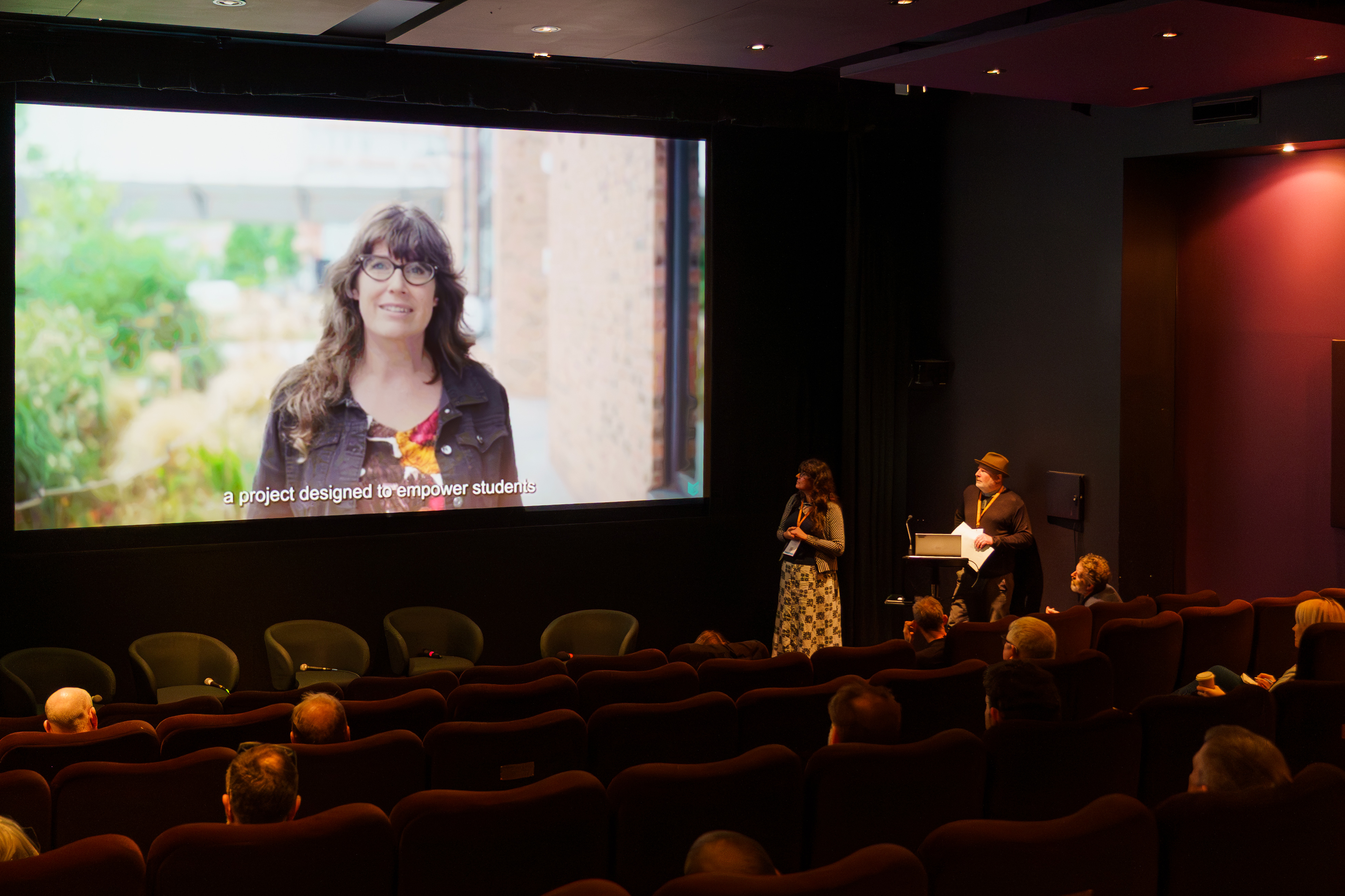 Liz is shown on a cinema screen as Matt and Liz deliver their talk to an audience.