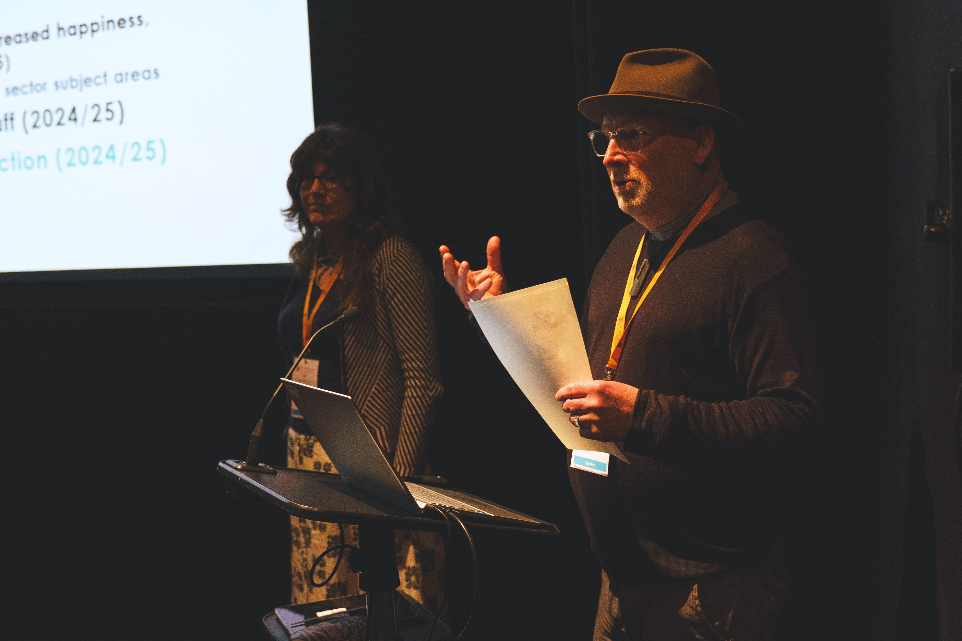 Matt is in the foreground, holding a piece of paper as he talks. Liz is behind him with a corner of a screen behind them.