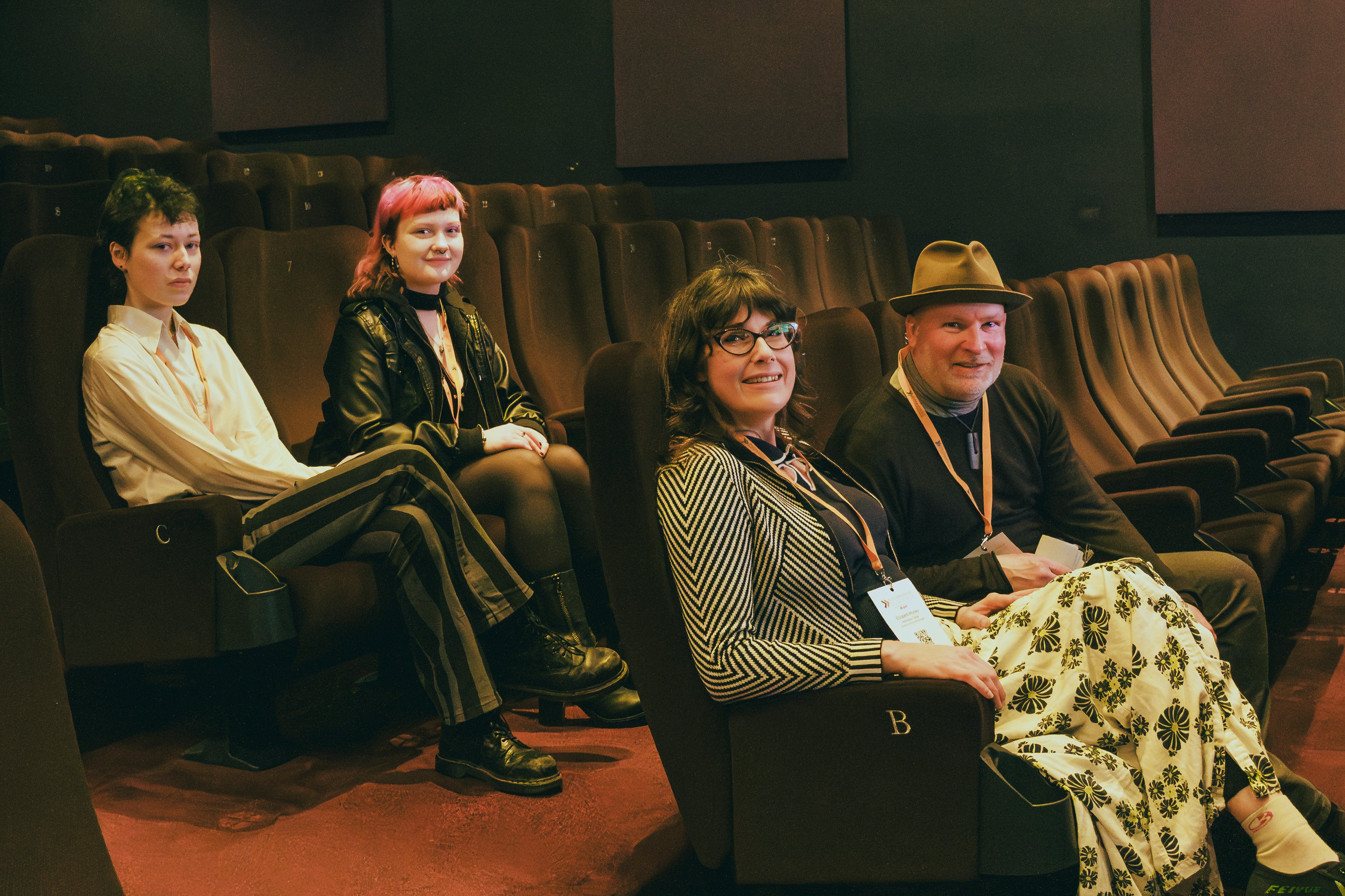 Matt and Liz sit on cinema chairs in a dimly lit screen with two students sat behind them. They all look and smile at the camera.