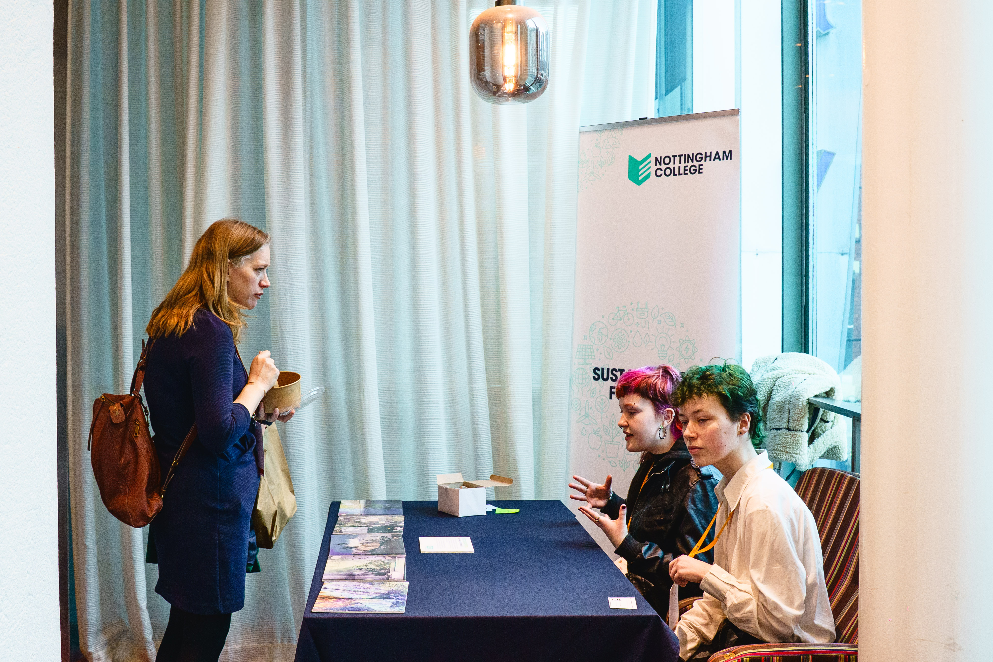A woman stands at a Nottingham College branded stall, clearly asking for information. Two students sit behind the staff responding to her.
