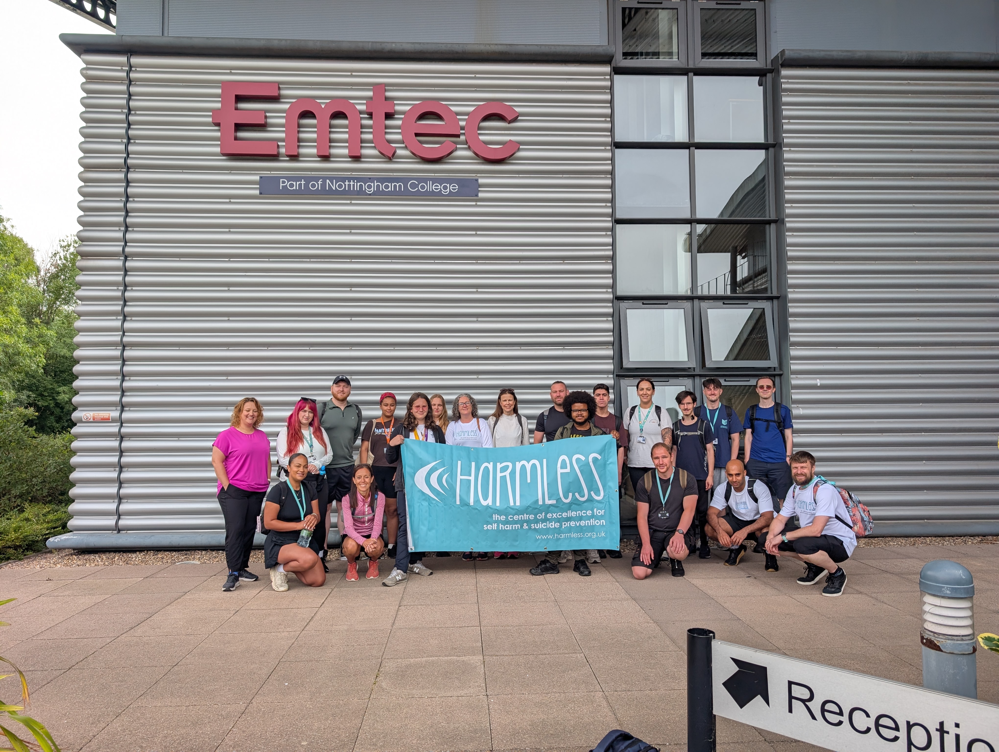 Staff and students outside EMTEC holding a banner that has the charity Harmless text written on it