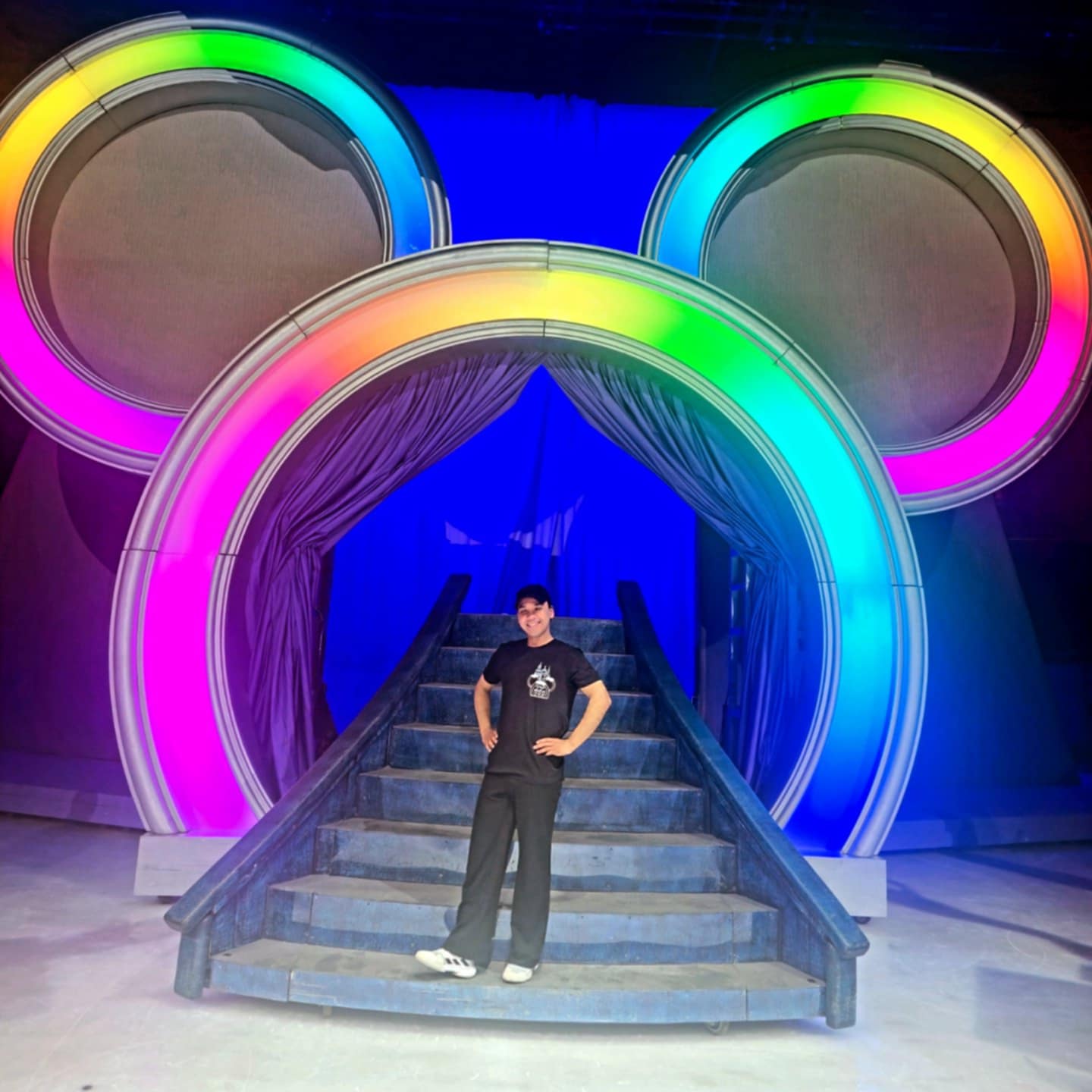 Hanyu poses on a small flight of stairs with a large colourful Mickey Mouse logo outline behind him.