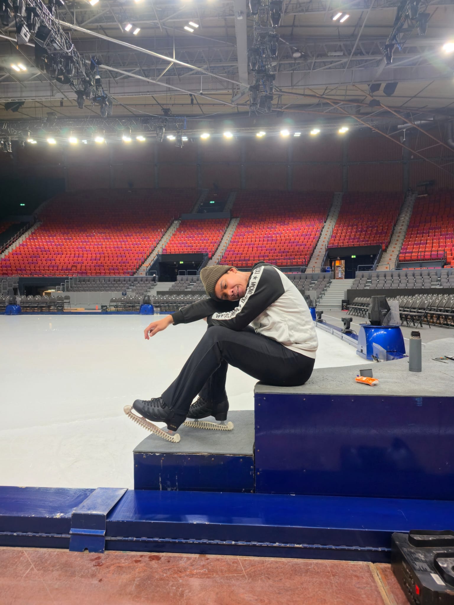 Hanyu poses sitting on some stairs at an ice rink with his ice skates on. The large arena can be seen behind him with red chairs.