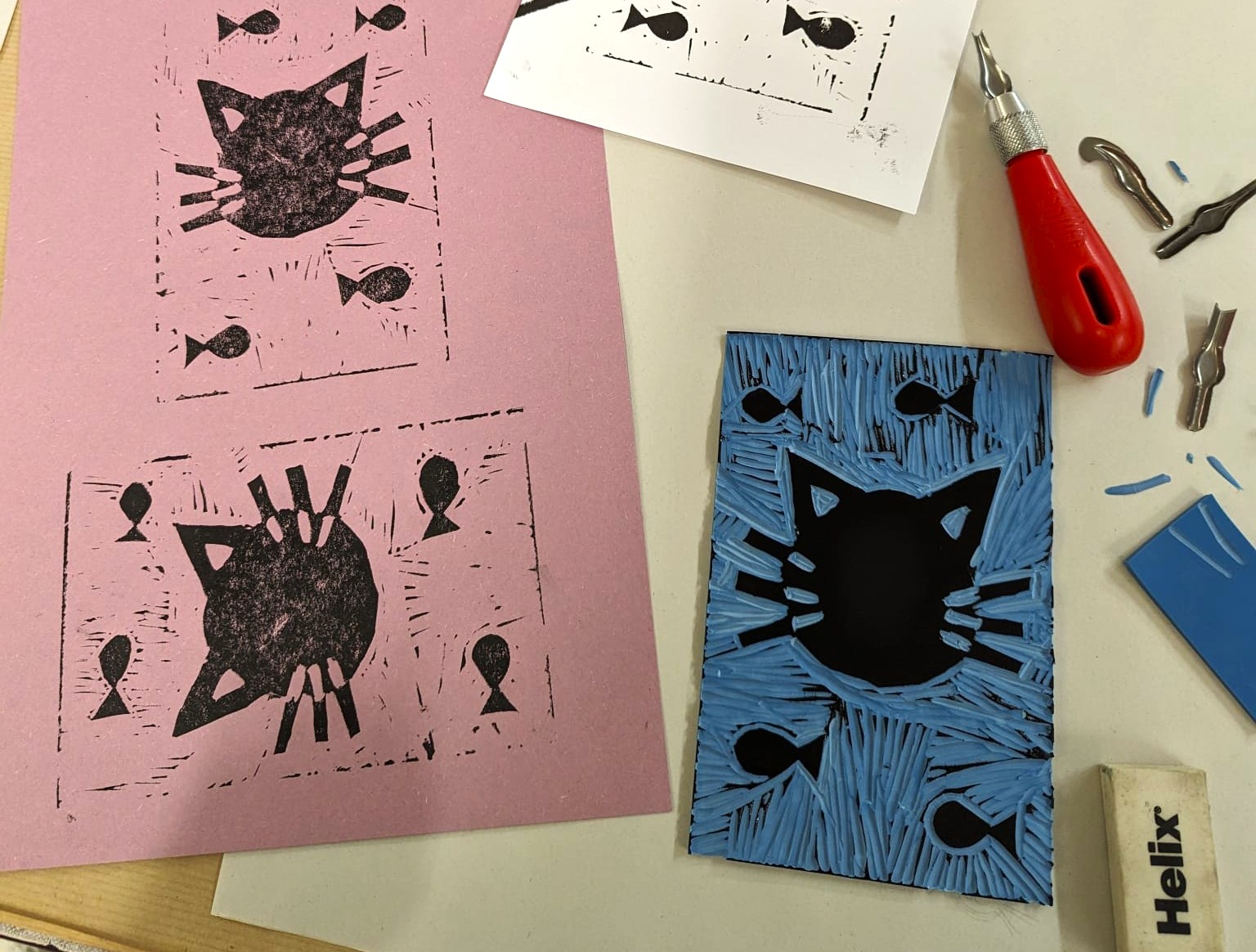 Photograph of a lino print in progress. Features two prints of a cat in black ink and the carved lino that produced them on a table with the lino cutting tool.