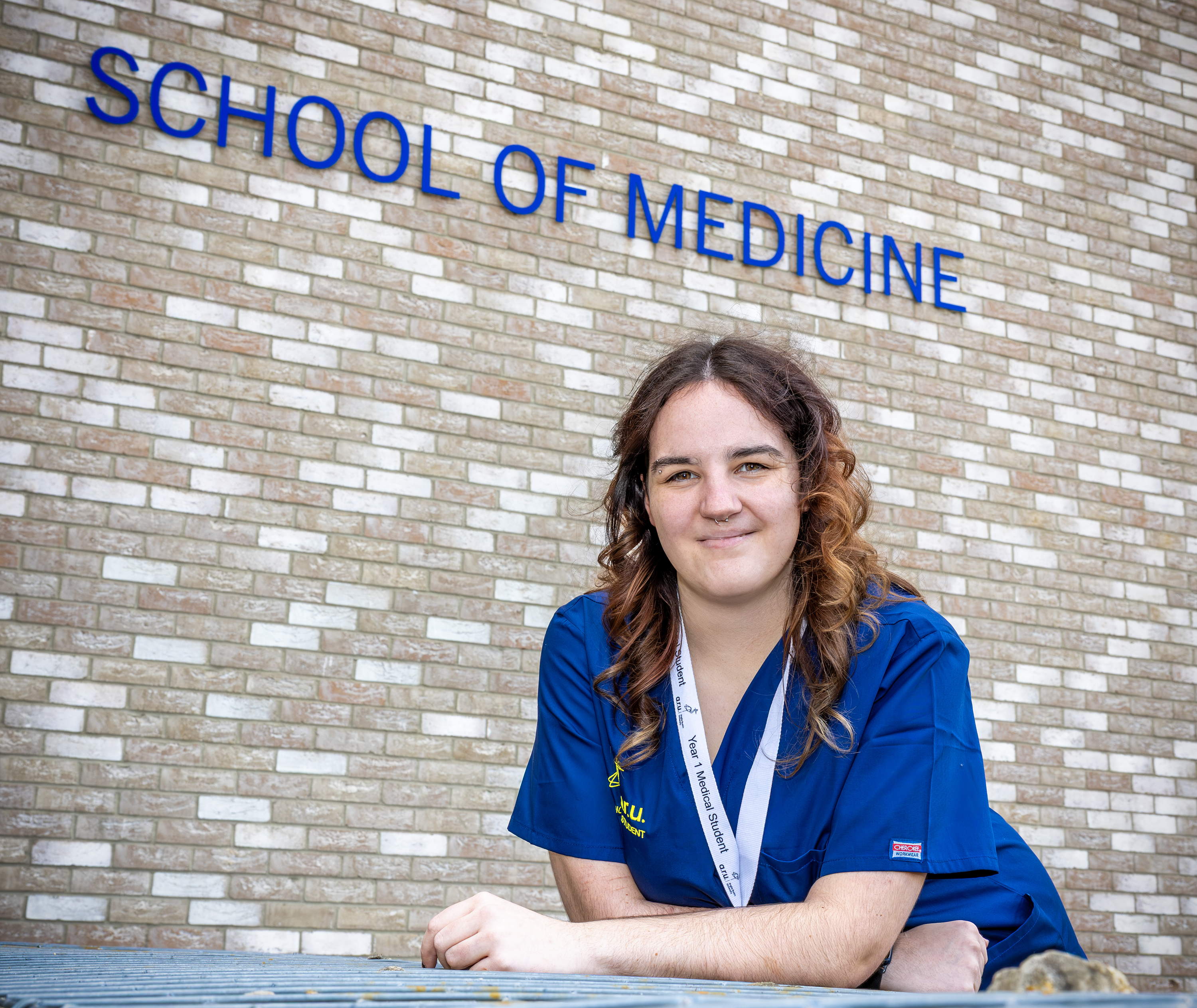 Photo of T Level student Katie wearing navy scrubs and lanyard in front of a sign that says 'School of Medicine'.