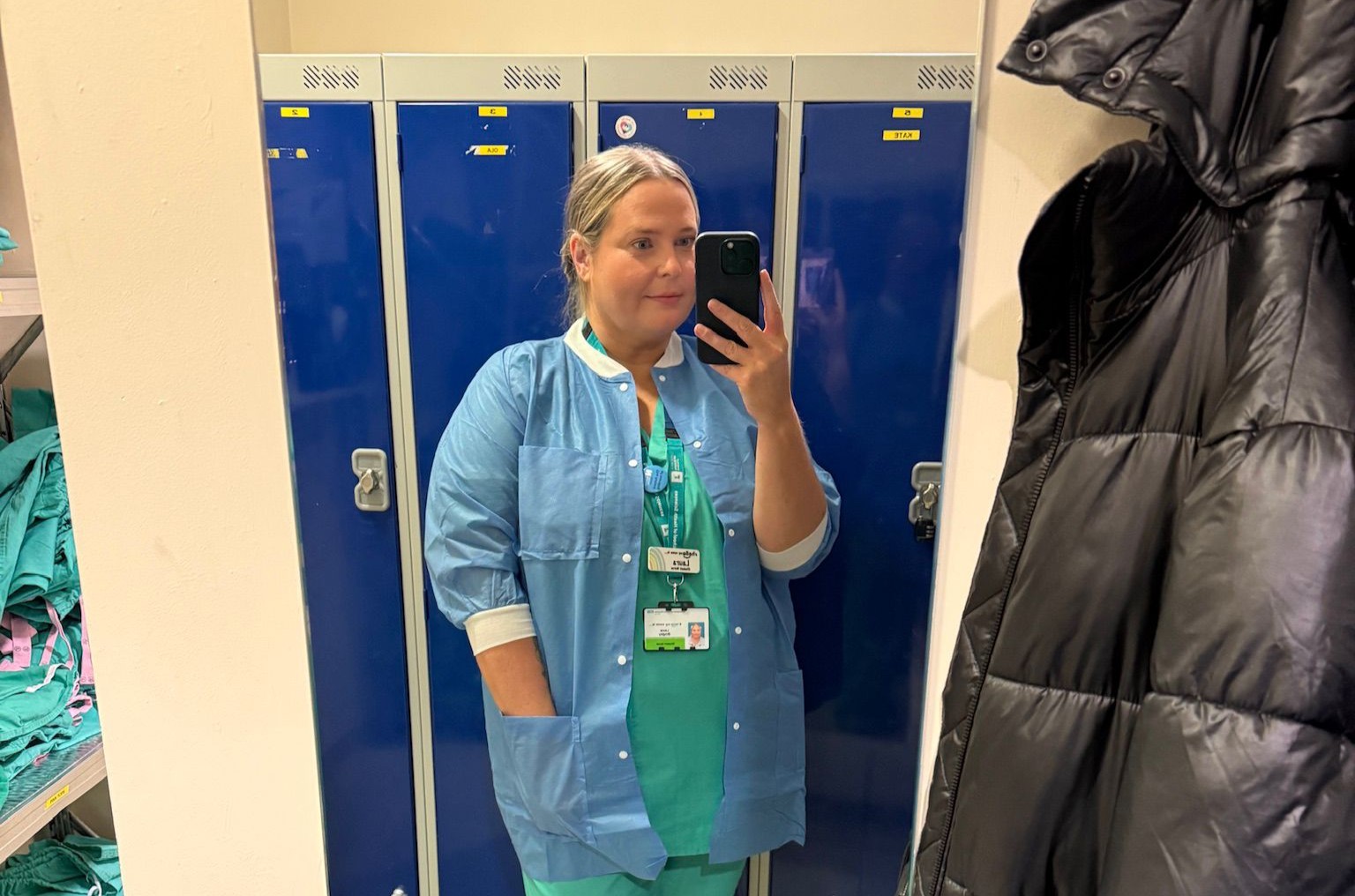Laura takes a photograph on her phone in the mirror of herself standing in front of blue lockers in her nurse scrubs.