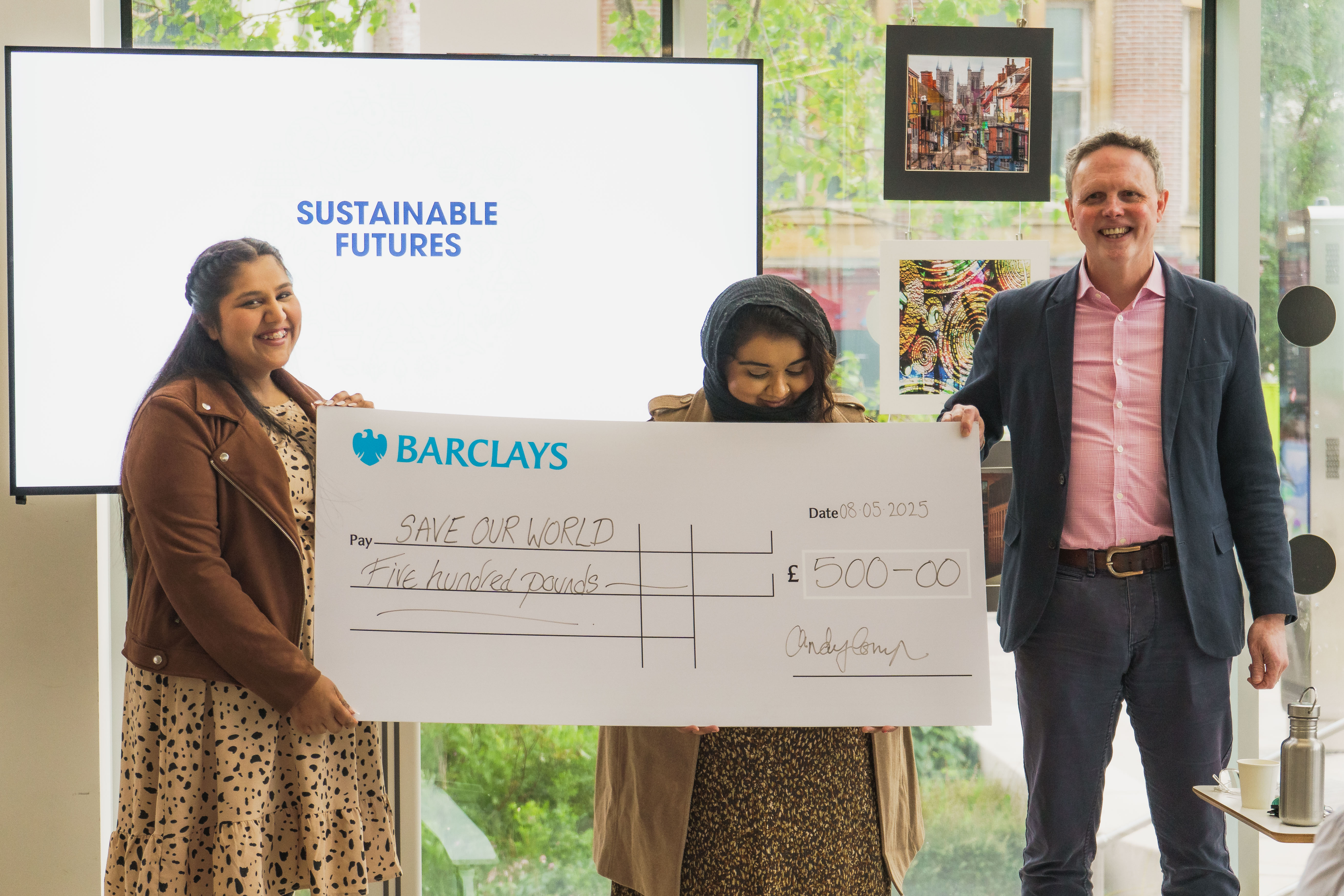 Two students smiling with a member of staff and the cheque they were awarded