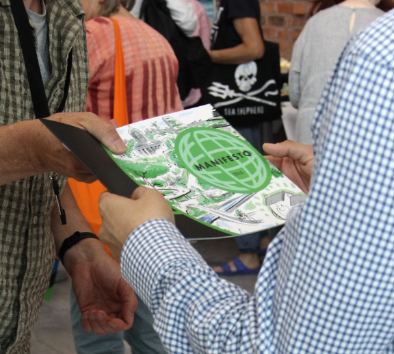 Two people in the foreground hold and peruse a leaflet with the word 'manifesto' on the front. People gather in the background. No face can be seen.