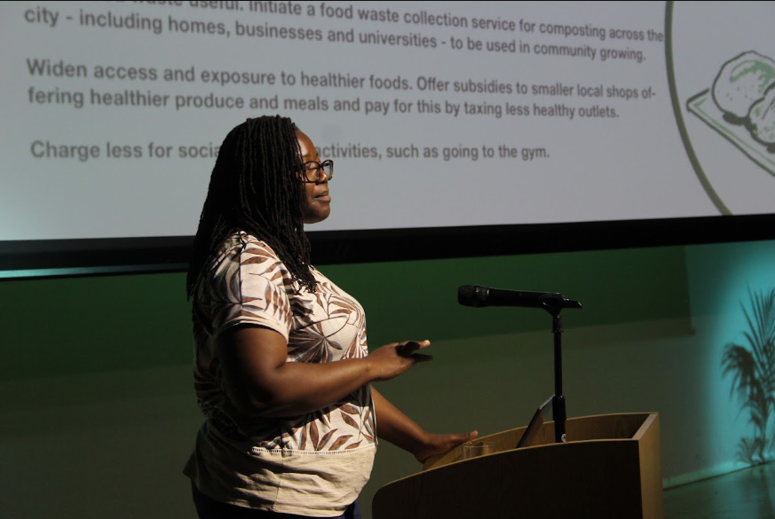 A photo of a woman speaking into a microphone in front of a presentation board displaying out of focus text.