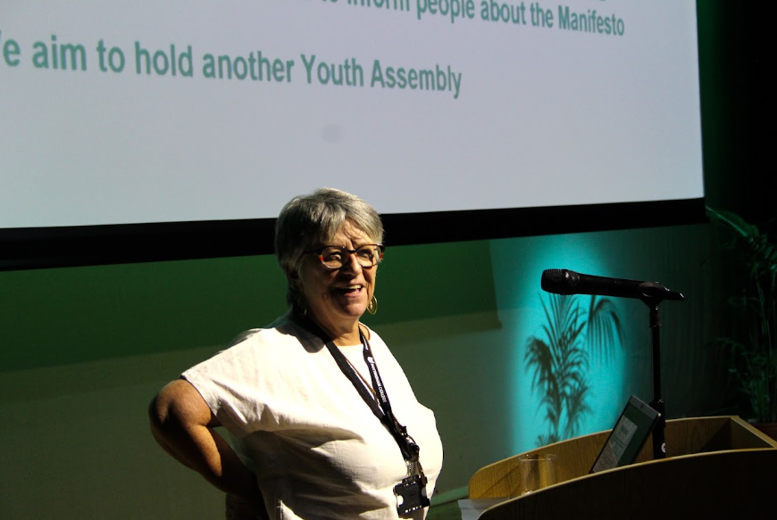 A photo of a lady wearing a white t-shirt smiling and talking into a microphone with a presentation behind her.