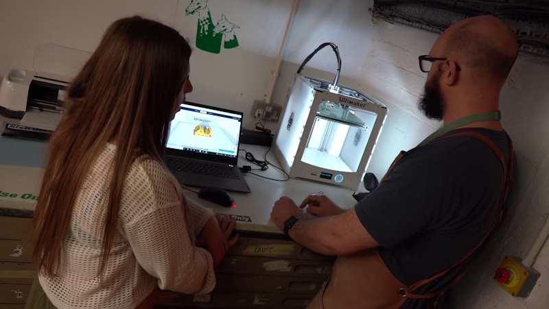 A student watching a lecturer operating a 3D printing machine with a laptop.