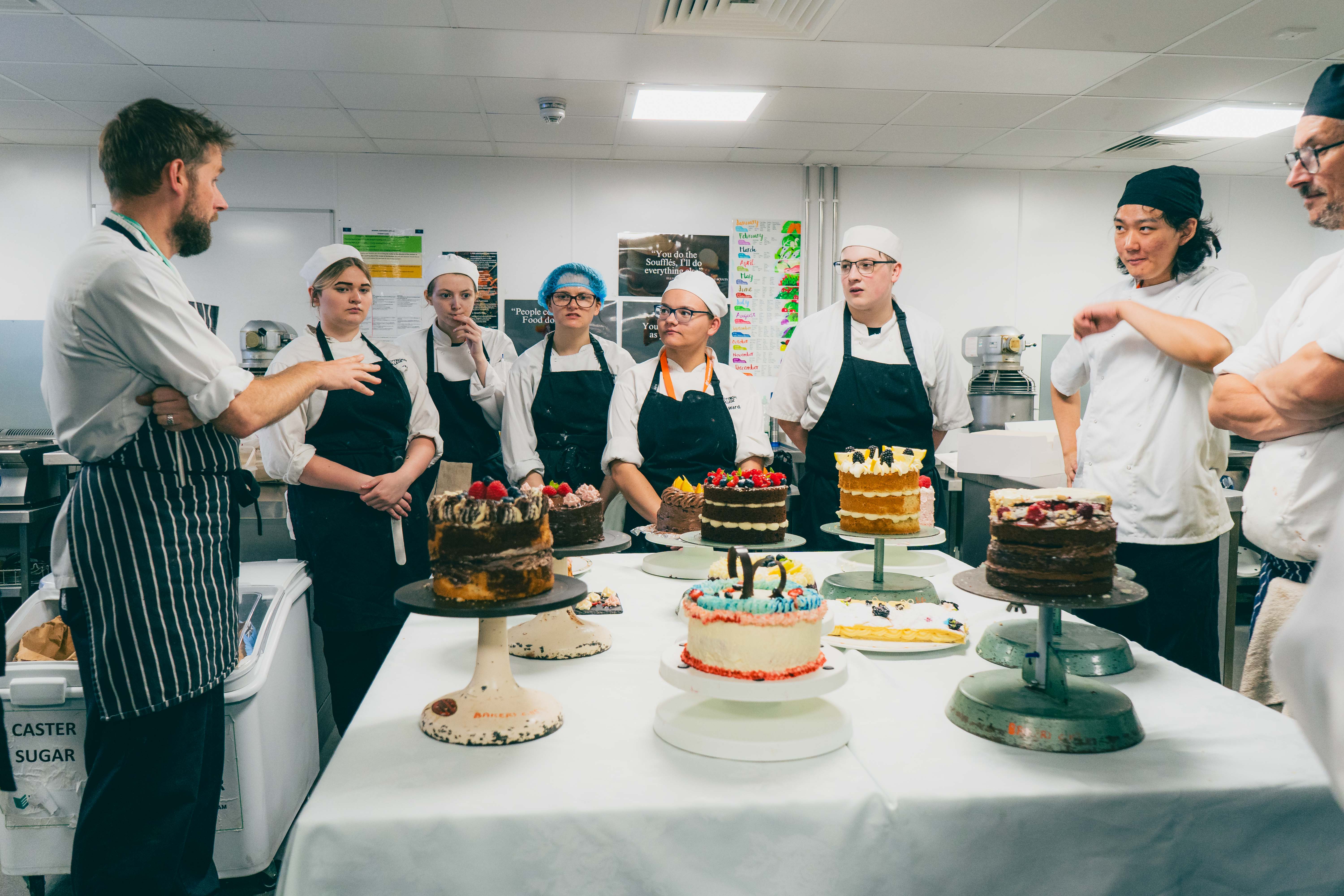 Students stood around a table with various cakes on listening to their tutor
