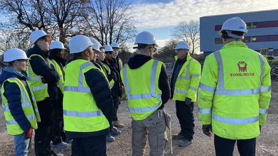 A group of students on a building site with high vis jackets and hard hats.