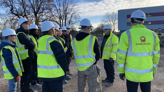A group of students on a building site with high vis jackets and hard hats.
