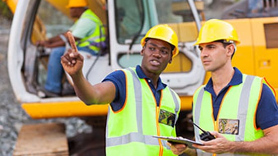 Two construction site workers talking in front of a mechanical digger.