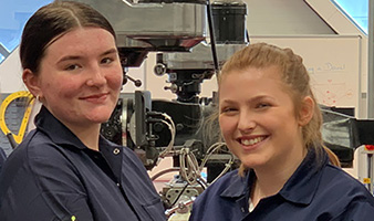 Two female engineering students smiling in front of engineering equipment