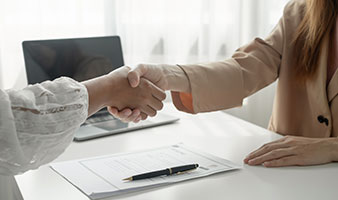Two people shaking hands over a desk with a laptop, pen and paper on the desk.