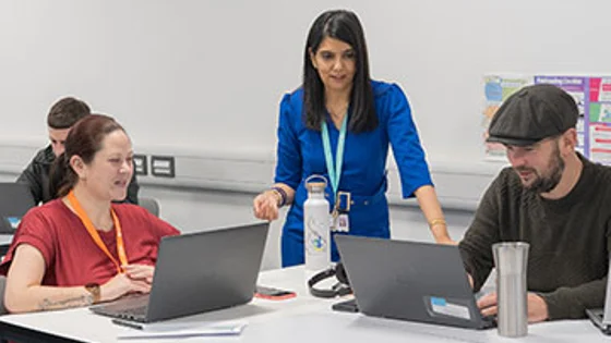 Two students sitting at a desk with laptops and a lecturer standing behind them.