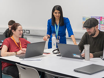 Two students sitting at a desk with laptops and a lecturer standing behind them.