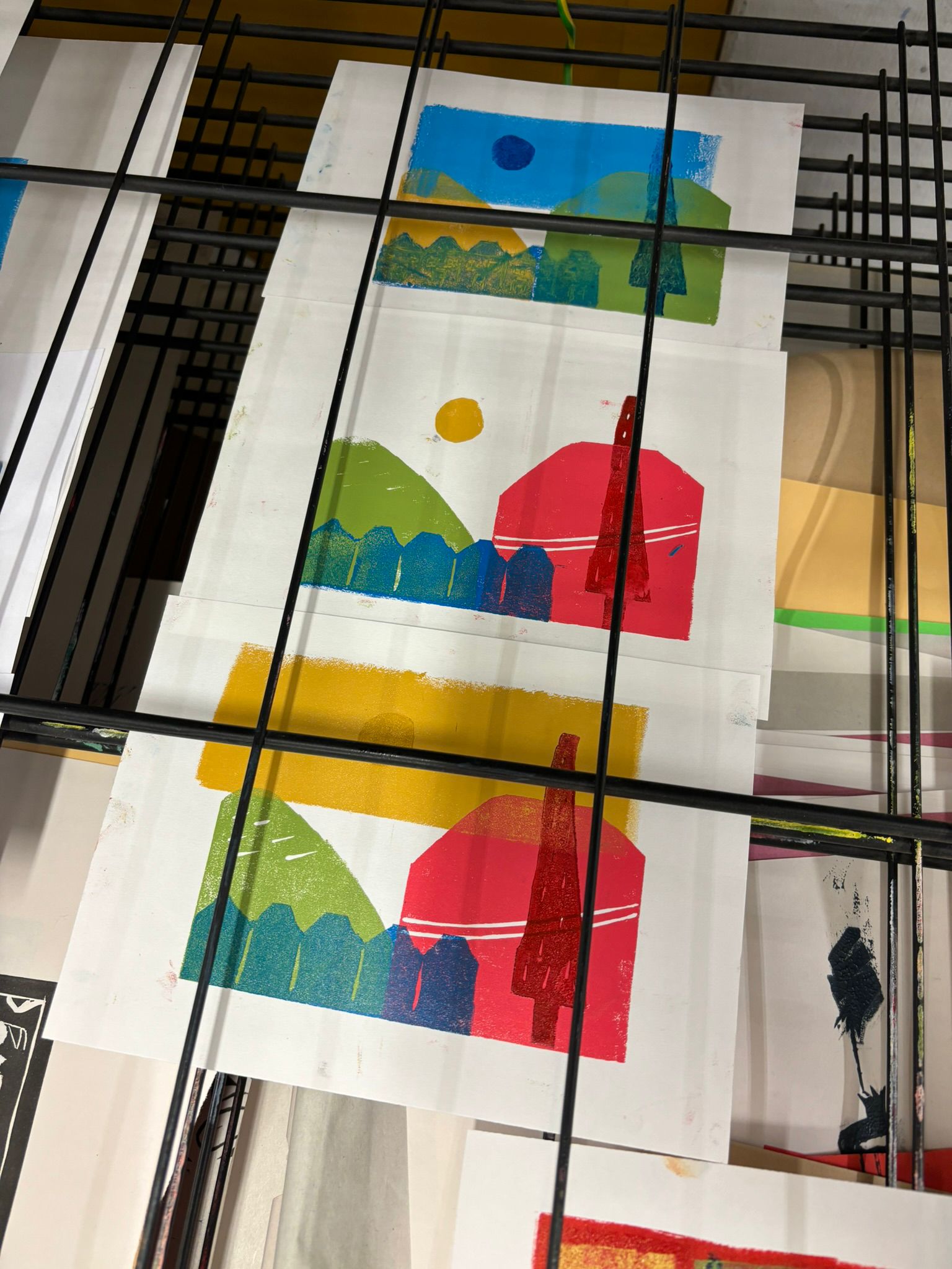 Image of three colourful lino prints on a drying rack. The prints are abstract shapes creating a landscape.