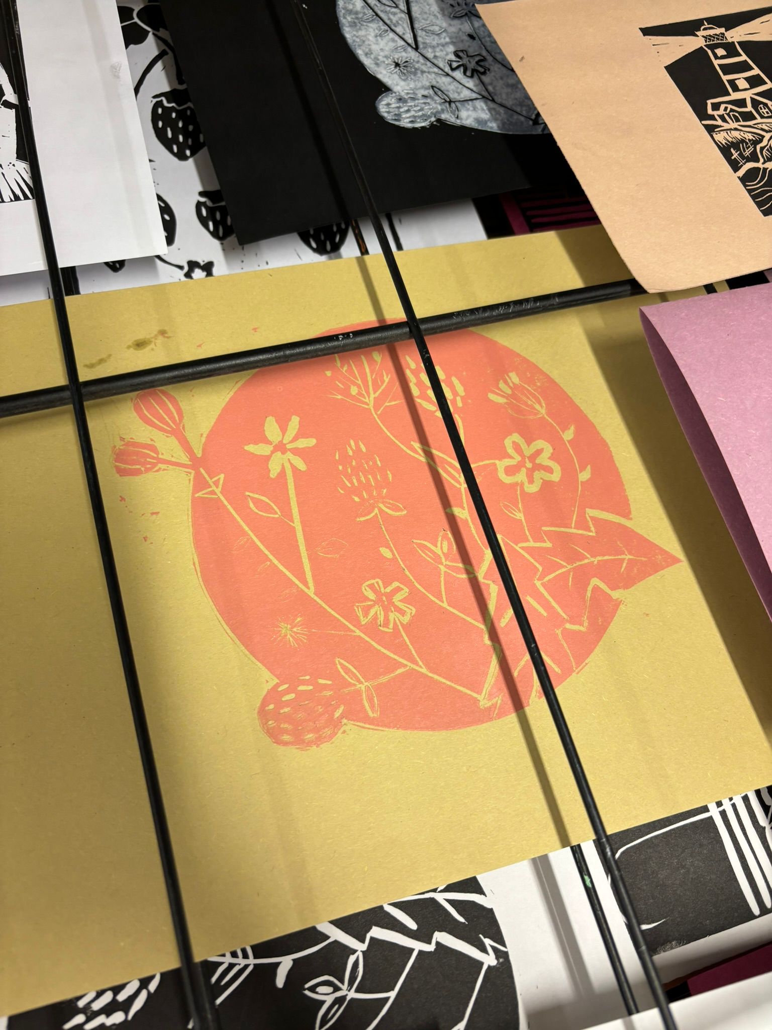 Image of a pink lino print of flowers on a yellow piece of paper, sitting on a drying rack.