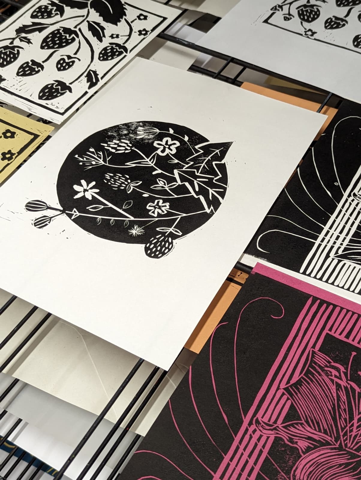 A photo of a few lino prints on a drying rack. The central print is by Lindsey and features a black ink image of flowers on a circle background.